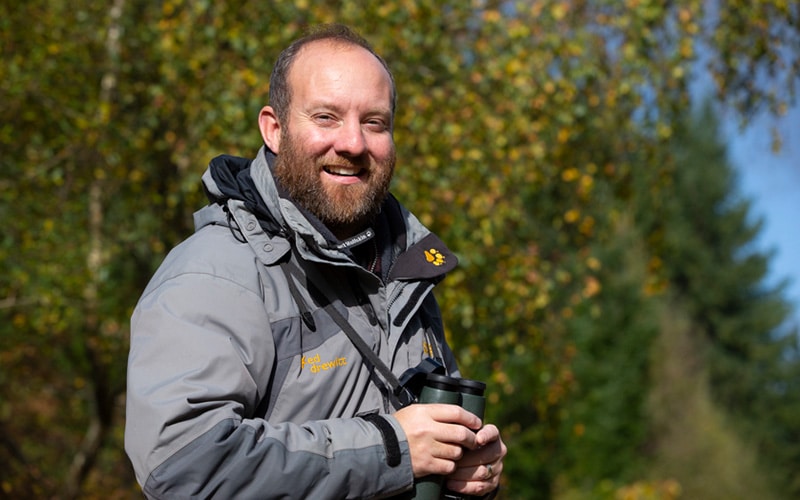 Ed Drewitt, naturalist with a pair of binoculars with a woodland behind him.
